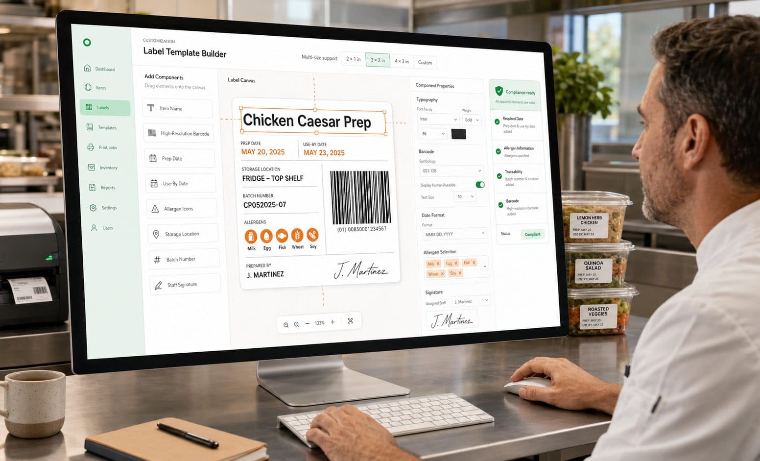 A chef at a stainless-steel kitchen bench works in the label template builder on a desktop monitor, dragging item name, prep date, use-by date, storage location, batch number, allergen icons and a staff signature onto a food label, with printed labels on prepped containers beside him.