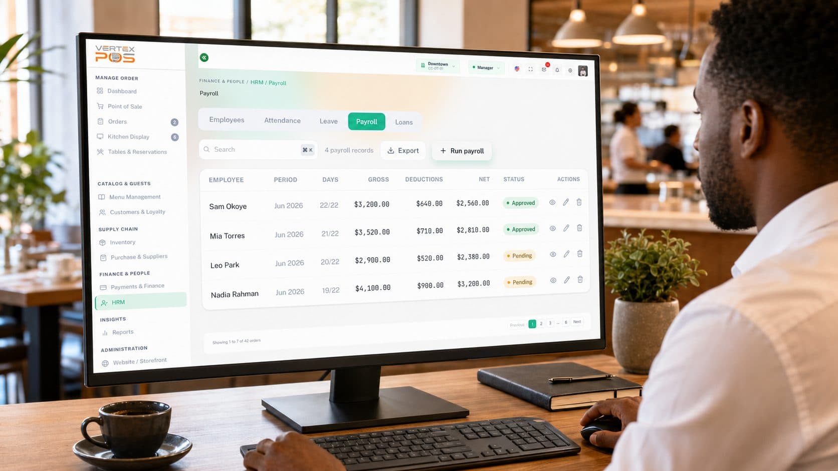 A manager reviewing the Vertex POS payroll dashboard on a desktop monitor in a café. The HRM workspace sits on the Payroll tab, next to Employees, Attendance, Leave and Loans, above a sample pay-run table listing each employee's pay period, days worked, gross pay, deductions, net pay and an Approved or Pending approval status.