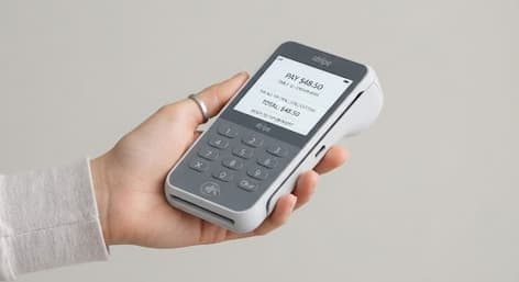 Handheld chip-and-PIN card reader held in one hand, its screen prompting the customer to pay $48.50