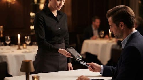 A server holds out a handheld card terminal so a seated guest can pay at the table in a candlelit dining room