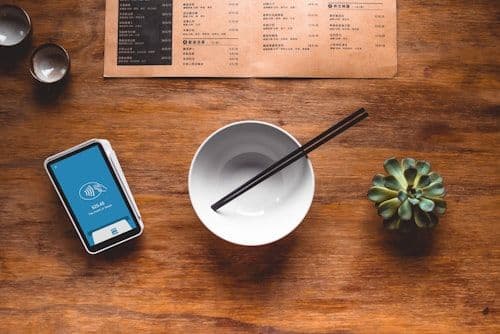 Handheld payment terminal resting on a restaurant table beside a bowl and chopsticks