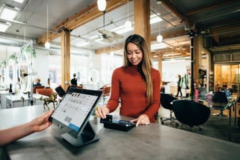 Tablet POS on a counter stand in a shop while an assistant helps a customer pay at a card reader.