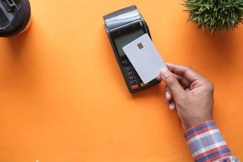 Compact card terminal with a receipt printer on an orange desk, a hand holding a chip card beside it.