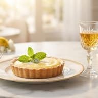 A small lemon tart topped with cream and a mint sprig on a rimmed plate, with a stemmed glass of golden dessert wine behind it.