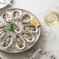 A round platter of freshly shucked oysters on crushed ice with a lemon wedge and dill, next to a glass of pale white wine on a marble table.