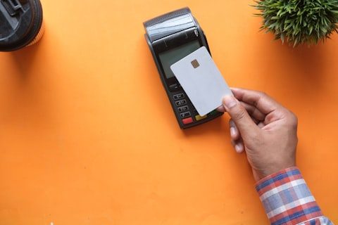 Compact card terminal with a receipt printer on an orange desk, a hand holding a chip card beside it.