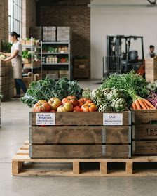 Wholesale produce warehouse: a staff member restocks shelving while crates of tomatoes, kale, artichokes and carrots wait on a pallet for despatch